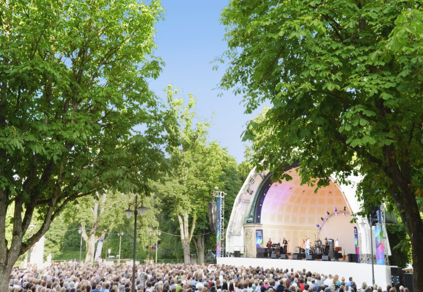 Publikum vor Open Air Bühne umringt von Bäumen bei blauem Himmel