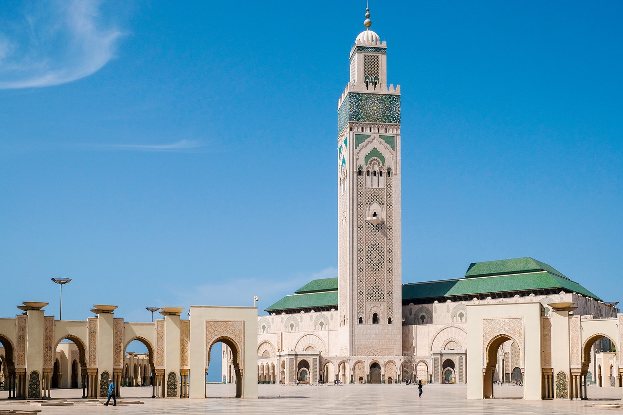 weiße Moschee mit grünem Dach und hohem Turm in der Mitte und vielen Torbögen vor blauem Himmel