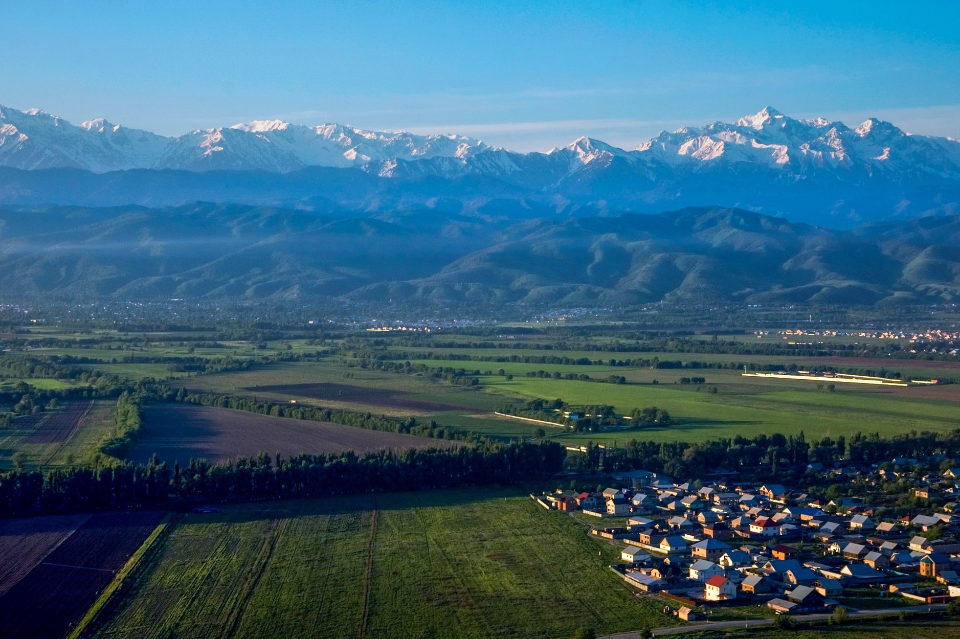 Blick über grüne Wiesen und Dorf auf Gebirgskette der Almaty Region