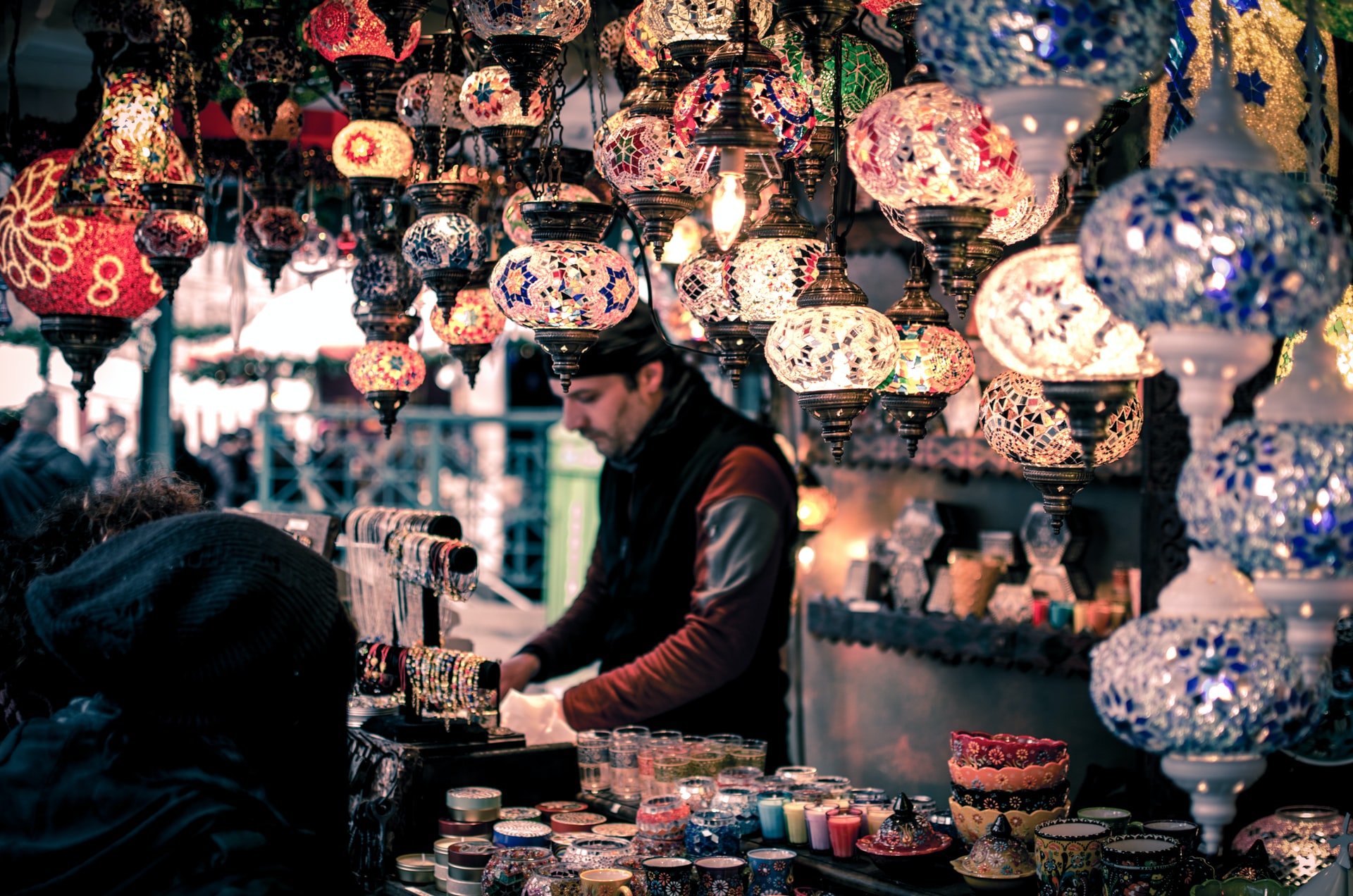 a man stands behind the counter of his market stall and above him hang colorful mosaic lamps