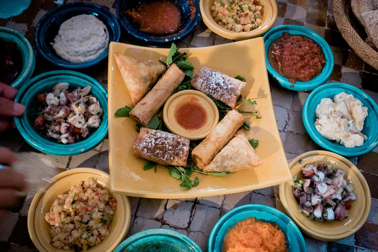 round yellow and blue plates filled with Moroccan food stand in a circle around a yellow square plate on which food is also placed