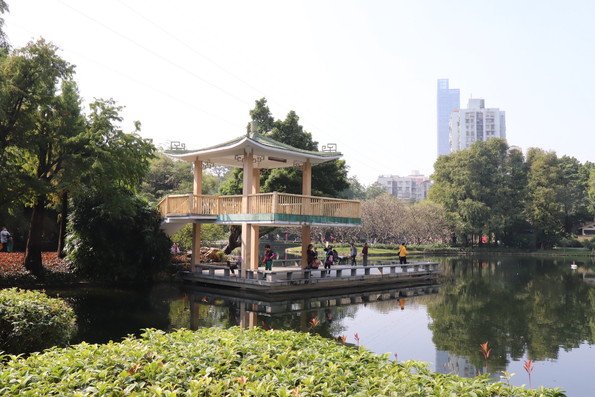 Chinese pavilion in park surrounded by trees and a pond in front of it, in the background you can see some skyscrapers