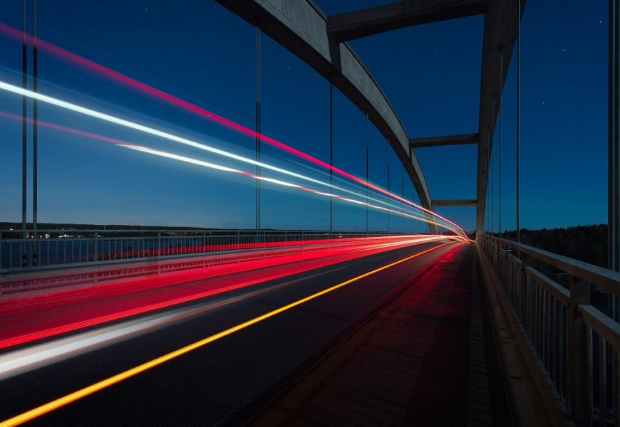 Das Foto wurde aus der Perspektive einer Person aufgenommen, die auf einer Brück bei Nacht steht und zeigt die Brückenpfosten und die Fahrbahn. Helle Lichtblitze auf der Fahrbahn deuten vorbeifahrende Fahrzeuge an.